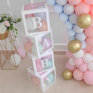 May include: White boxes spelling "BABY" with pastel-coloured balloons inside. The boxes are stacked, with the letters in white. A balloon arch and gold balloons are in the background. The theme is a baby shower or gender reveal.