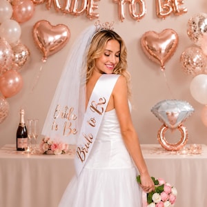 Könnte beinhalten: Ein weißer Brautschleier mit roségoldenem Bride to Be Schriftzug, eine weiße Satinschärpe mit roségoldener Bride to Be Aufschrift und ein roségoldener Metallhaarreif mit dem Wort Bride, getragen zu einem weißen Kleid und einem Rosenstrauß.