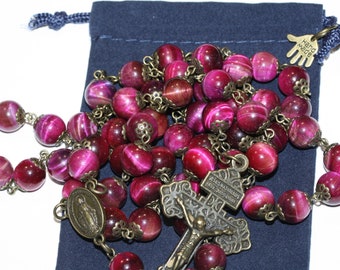 May include: A rosary with large, round, magenta tiger eye beads and antique bronze-colored metal accents. The rosary includes a crucifix and a medal of the Virgin Mary. A navy blue pouch is in the background.