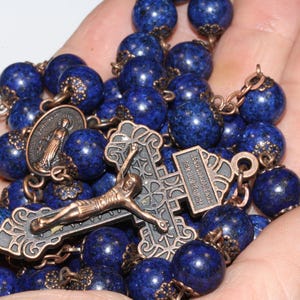 May include: A rosary with deep blue, spherical beads. The beads are strung together with metal chain and connectors. A detailed metal crucifix and a medallion are also present. The rosary is held in a hand.