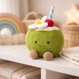Op de afbeelding: Een groene pluche kokosnootspeelgoed met een lachend gezicht, kleine bruine voetjes en een decoratieve bovenkant. De bovenkant heeft een witte bloem, een rode kers, een gele pompon en een blauw-wit gestreept rietje. Het speelgoed staat op een witte plank.