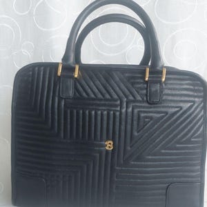 May include: Black leather handbag with geometric stitching and gold-tone hardware. Features two top handles and a rectangular shape. The bag has a gold-colored emblem on the front. The bag is sitting on a white surface.