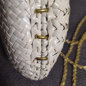 May include: A cream-colored woven handbag featuring a glossy finish and a structured design. The bag is accented with three horizontal gold-toned metal bars along the center seam and includes a delicate gold-toned chain strap for carrying.