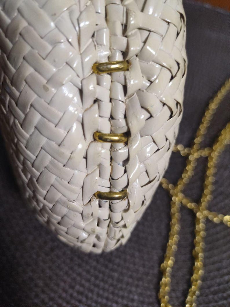 May include: A cream-colored woven handbag featuring a glossy finish and a structured design. The bag is accented with three horizontal gold-toned metal bars along the center seam and includes a delicate gold-toned chain strap for carrying.