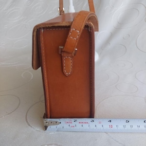 May include: A rectangular, brown leather crossbody bag with a flap closure and a shoulder strap. The bag is approximately 5 inches tall and 3 inches wide. The stitching is visible in white thread, and the bag has a buckle closure.