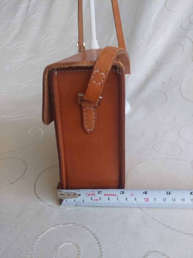 May include: A rectangular, brown leather crossbody bag with a flap closure and a shoulder strap. The bag is approximately 5 inches tall and 3 inches wide. The stitching is visible in white thread, and the bag has a buckle closure.