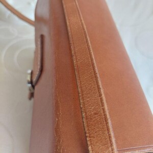 May include: A brown leather handbag with a shoulder strap. The bag has a rectangular shape with a flap closure and a visible buckle. The leather shows signs of wear, adding to its vintage appeal. The strap is attached to the bag's sides.