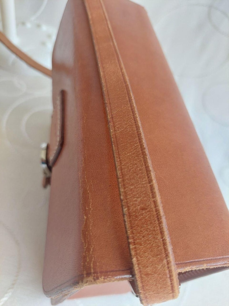 May include: A brown leather handbag with a shoulder strap. The bag has a rectangular shape with a flap closure and a visible buckle. The leather shows signs of wear, adding to its vintage appeal. The strap is attached to the bag's sides.