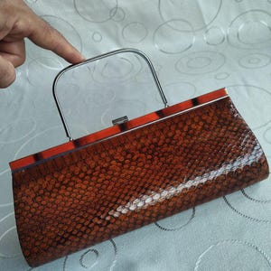 May include: A rectangular clutch bag featuring a brown and black snakeskin-patterned exterior with a glossy finish. The bag includes a silver-toned metal top handle and a matching silver-toned metal frame along the top edge with a small clasp closure.