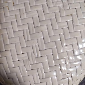 May include: A close-up view of a white woven surface featuring a repeating herringbone pattern. The material consists of flat, smooth strips interlaced in a tight, diagonal configuration, creating a consistent texture across the entire visible area.