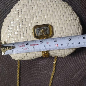 May include: A cream-colored woven handbag with a gold-tone metal clasp featuring the letters "FG". A measuring tape placed across the front shows the bag is approximately 6 inches wide. It includes a thin gold-tone metal chain shoulder strap.