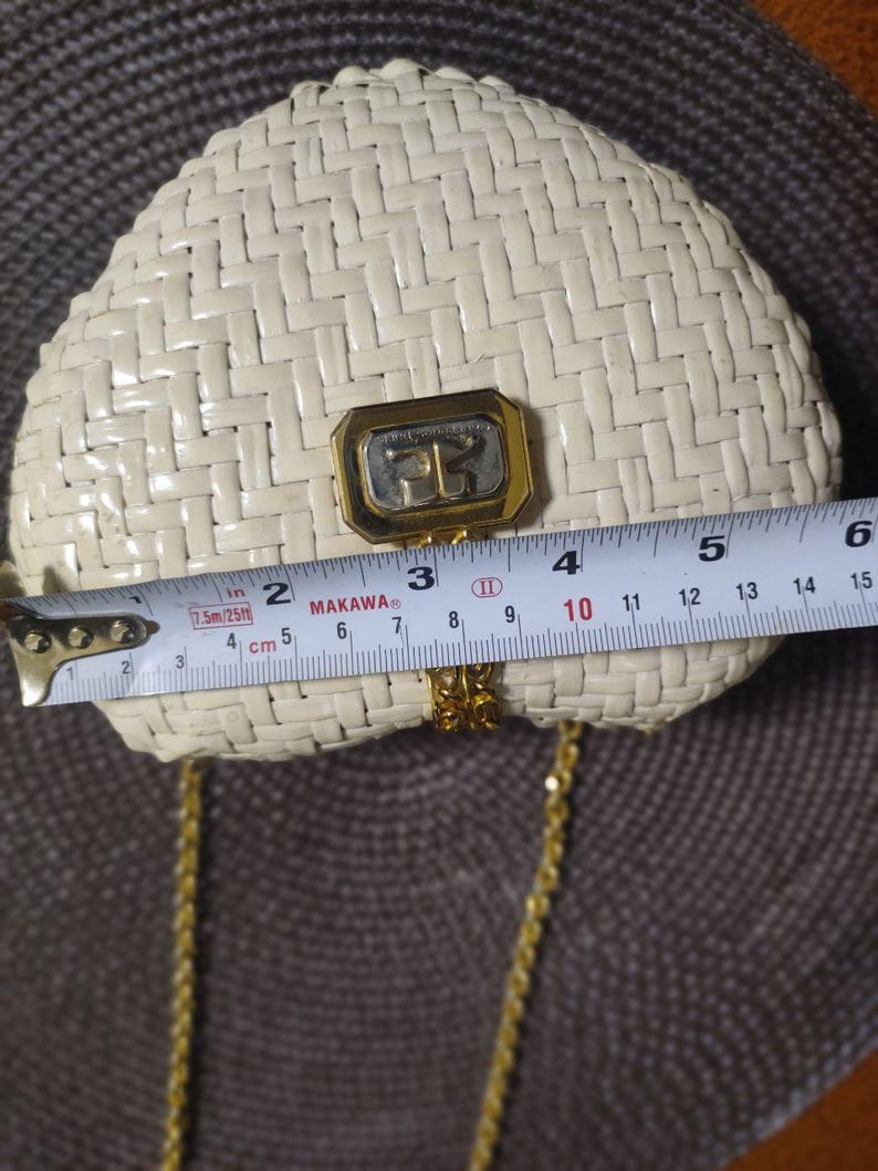 May include: A cream-colored woven handbag with a gold-tone metal clasp featuring the letters "FG". A measuring tape placed across the front shows the bag is approximately 6 inches wide. It includes a thin gold-tone metal chain shoulder strap.