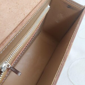 May include: Open view of a brown leather handbag with a rectangular shape. The interior is light brown with a silver zipper and white stitching. The bag has a small strap.
