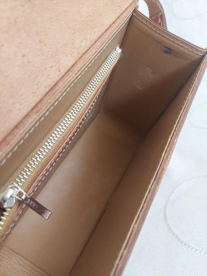 May include: Open view of a brown leather handbag with a rectangular shape. The interior is light brown with a silver zipper and white stitching. The bag has a small strap.