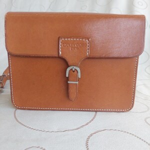 May include: A rectangular, tan leather Courreges purse with a flap closure and silver buckle. The purse has white stitching around the edges and a shoulder strap. The Courreges logo is embossed on the front.