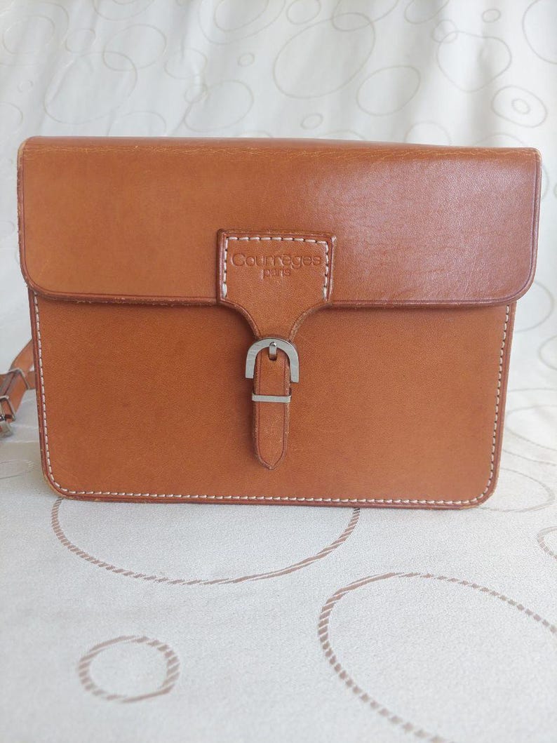 May include: A rectangular, tan leather Courreges purse with a flap closure and silver buckle. The purse has white stitching around the edges and a shoulder strap. The Courreges logo is embossed on the front.