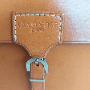 May include: Close-up of a tan leather bag with a silver buckle. The bag features a rectangular leather patch with the brand name "Courr&egrave;ges paris" stitched on it. The leather has a smooth texture.