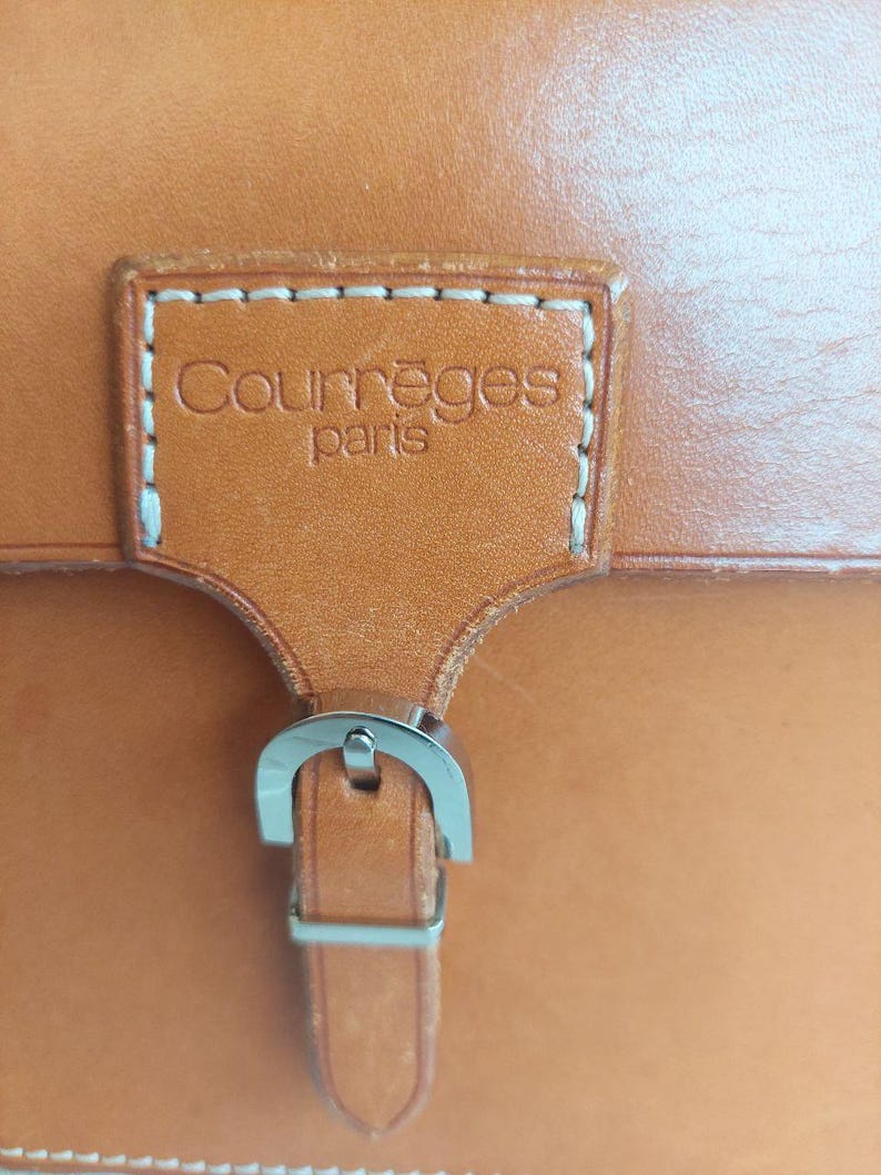 May include: Close-up of a tan leather bag with a silver buckle. The bag features a rectangular leather patch with the brand name "Courr&egrave;ges paris" stitched on it. The leather has a smooth texture.