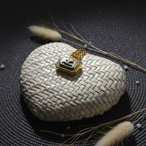 May include: A gold-toned pendant necklace featuring a rectangular charm with a black center inlay and a metallic logo emblem. The pendant is attached to a delicate gold-toned chain, showcasing a polished finish and a structured geometric design.