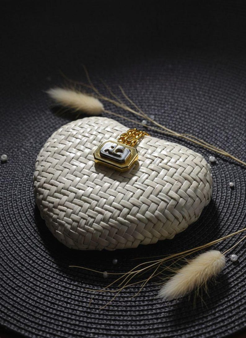 May include: A gold-toned pendant necklace featuring a rectangular charm with a black center inlay and a metallic logo emblem. The pendant is attached to a delicate gold-toned chain, showcasing a polished finish and a structured geometric design.