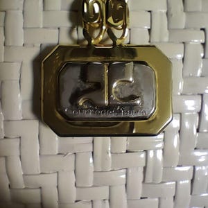 May include: A white woven handbag featuring a gold-tone metal plaque with a silver-tone center. The plaque displays the Courr&egrave;ges Paris logo and text. A gold-tone chain link strap is attached to the top of the rectangular metal hardware.