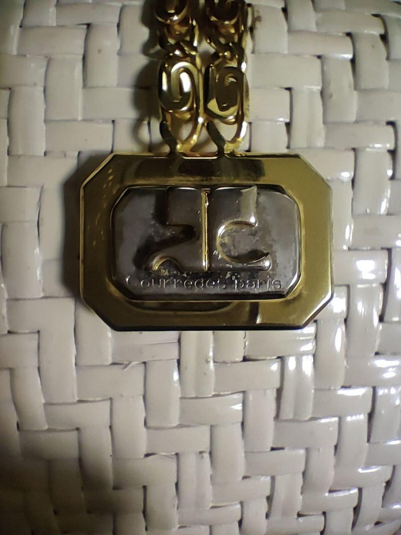 May include: A white woven handbag featuring a gold-tone metal plaque with a silver-tone center. The plaque displays the Courr&egrave;ges Paris logo and text. A gold-tone chain link strap is attached to the top of the rectangular metal hardware.