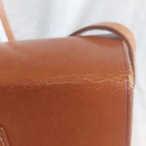 May include: Close-up of a brown leather crossbody bag. The bag has a flap closure and a long strap. The leather shows signs of wear, with visible creases and imperfections. The bag's color is a warm, medium brown.