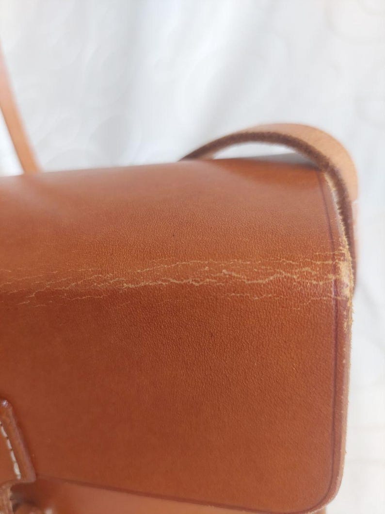 May include: Close-up of a brown leather crossbody bag. The bag has a flap closure and a long strap. The leather shows signs of wear, with visible creases and imperfections. The bag's color is a warm, medium brown.