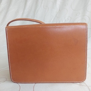 May include: A rectangular, tan leather handbag with a small handle. The bag features white stitching around the edges and a smooth, polished surface. The bag is designed with a flap closure.