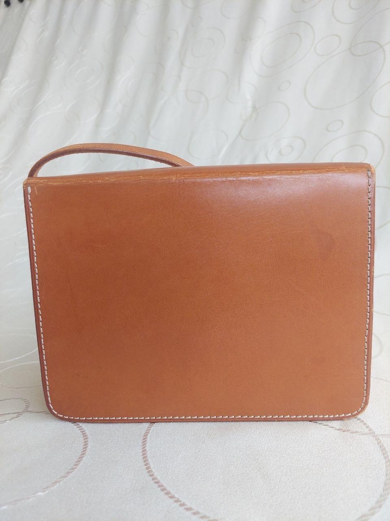 May include: A rectangular, tan leather handbag with a small handle. The bag features white stitching around the edges and a smooth, polished surface. The bag is designed with a flap closure.