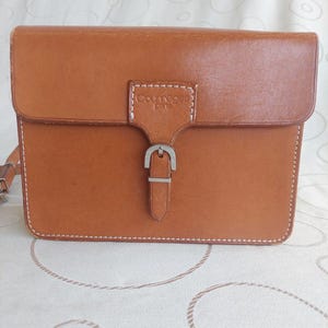 May include: A rectangular, tan leather Courreges purse with a flap closure and silver buckle. The purse has white stitching around the edges and a shoulder strap. The Courreges logo is embossed on the front.