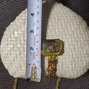 May include: A cream-colored woven handbag with a gold-tone metal clasp and a matching gold chain strap. A measuring tape is placed vertically across the center, showing the bag height is approximately 5.5 inches from the top to the bottom edge.