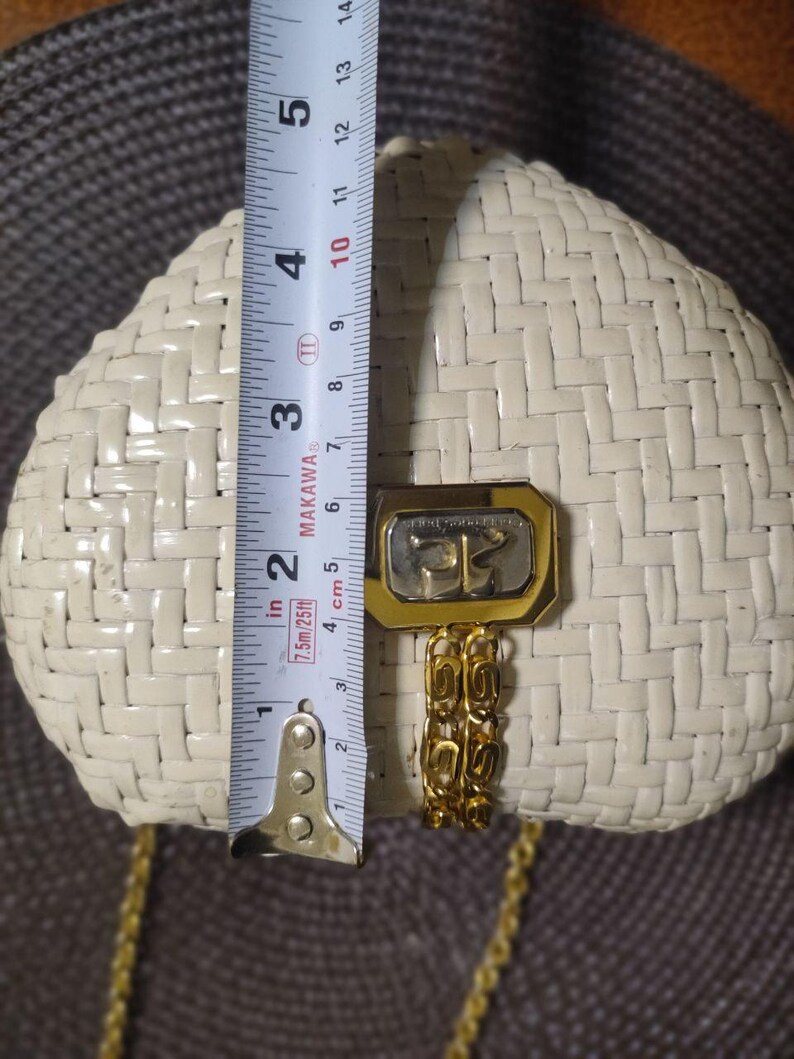 May include: A cream-colored woven handbag with a gold-tone metal clasp and a matching gold chain strap. A measuring tape is placed vertically across the center, showing the bag height is approximately 5.5 inches from the top to the bottom edge.