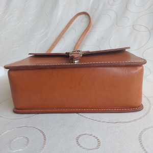 May include: A rectangular, brown leather handbag with a top flap and a buckle closure. The bag has a handle and is stitched with white thread. The bag is sitting on a white surface.