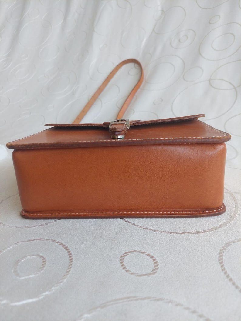 May include: A rectangular, brown leather handbag with a top flap and a buckle closure. The bag has a handle and is stitched with white thread. The bag is sitting on a white surface.