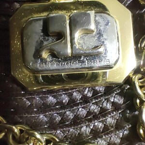 May include: A gold-toned metal pendant featuring a rectangular silver-toned center with the embossed logo and the text courreges paris. The pendant is attached to a thick gold-toned chain link necklace with a polished finish and reflective surface.