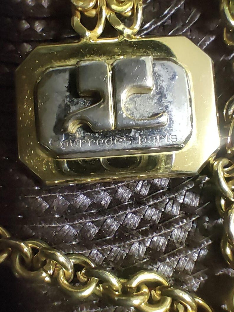 May include: A gold-toned metal pendant featuring a rectangular silver-toned center with the embossed logo and the text courreges paris. The pendant is attached to a thick gold-toned chain link necklace with a polished finish and reflective surface.