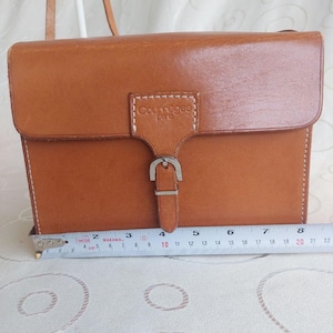 May include: A rectangular, brown leather crossbody bag with a flap closure and silver buckle. The bag has a shoulder strap and the text "Courreges paris" is visible. The bag measures approximately 8 inches wide.