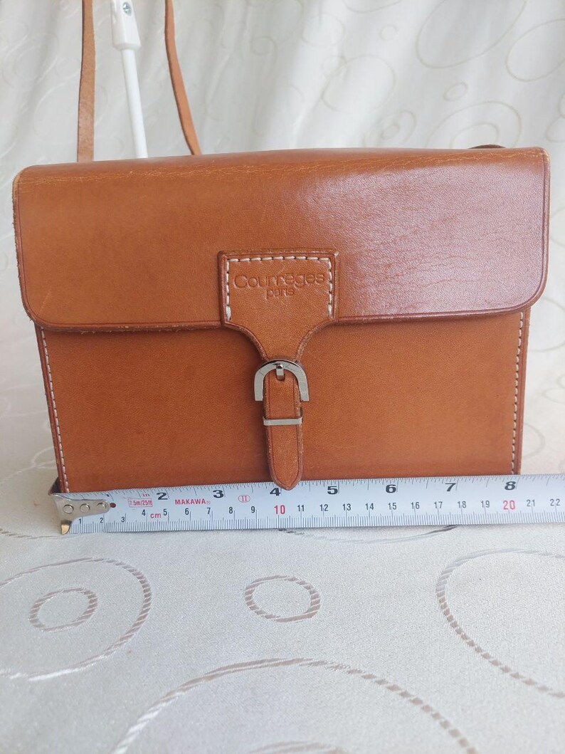 May include: A rectangular, brown leather crossbody bag with a flap closure and silver buckle. The bag has a shoulder strap and the text "Courreges paris" is visible. The bag measures approximately 8 inches wide.