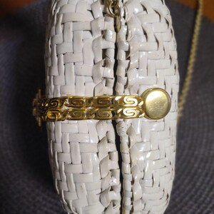 May include: A white woven clutch bag featuring a gold-tone metal chain strap and a decorative gold-tone metal side accent. The bag has a structured, rounded shape with a visible gap between the two halves, secured by a gold-tone metal clasp.