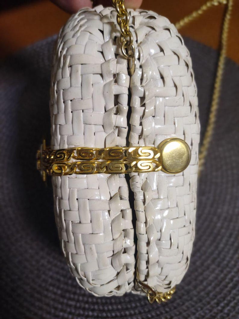 May include: A white woven clutch bag featuring a gold-tone metal chain strap and a decorative gold-tone metal side accent. The bag has a structured, rounded shape with a visible gap between the two halves, secured by a gold-tone metal clasp.