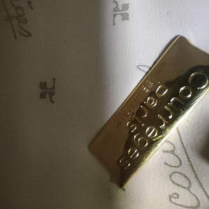 May include: A gold-toned rectangular metal plaque featuring the embossed text Courr&egrave;ges Paris and Made in Italy. The plaque is attached to a light-colored interior lining that displays a repeating pattern of the Courr&egrave;ges logo and script text.