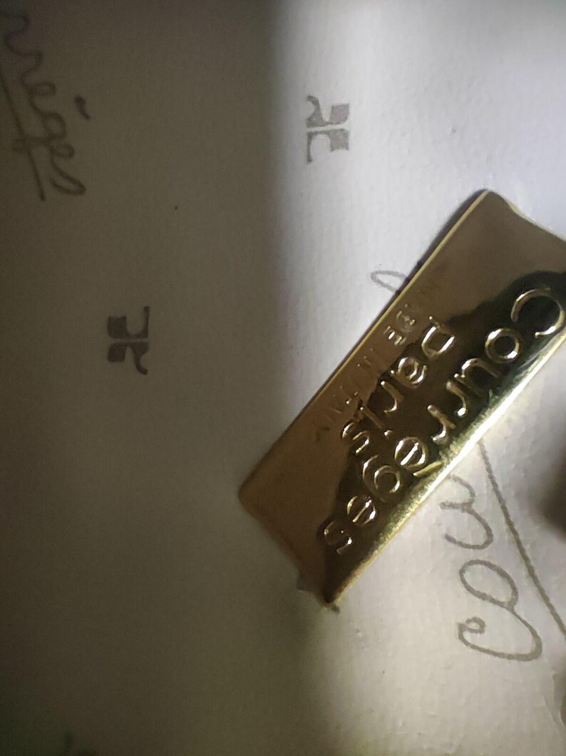 May include: A gold-toned rectangular metal plaque featuring the embossed text Courr&egrave;ges Paris and Made in Italy. The plaque is attached to a light-colored interior lining that displays a repeating pattern of the Courr&egrave;ges logo and script text.