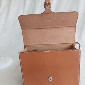 May include: Open, light brown leather handbag with a rectangular shape. The bag features white stitching, a top handle, and a metal clasp closure. The interior reveals a zippered pocket.