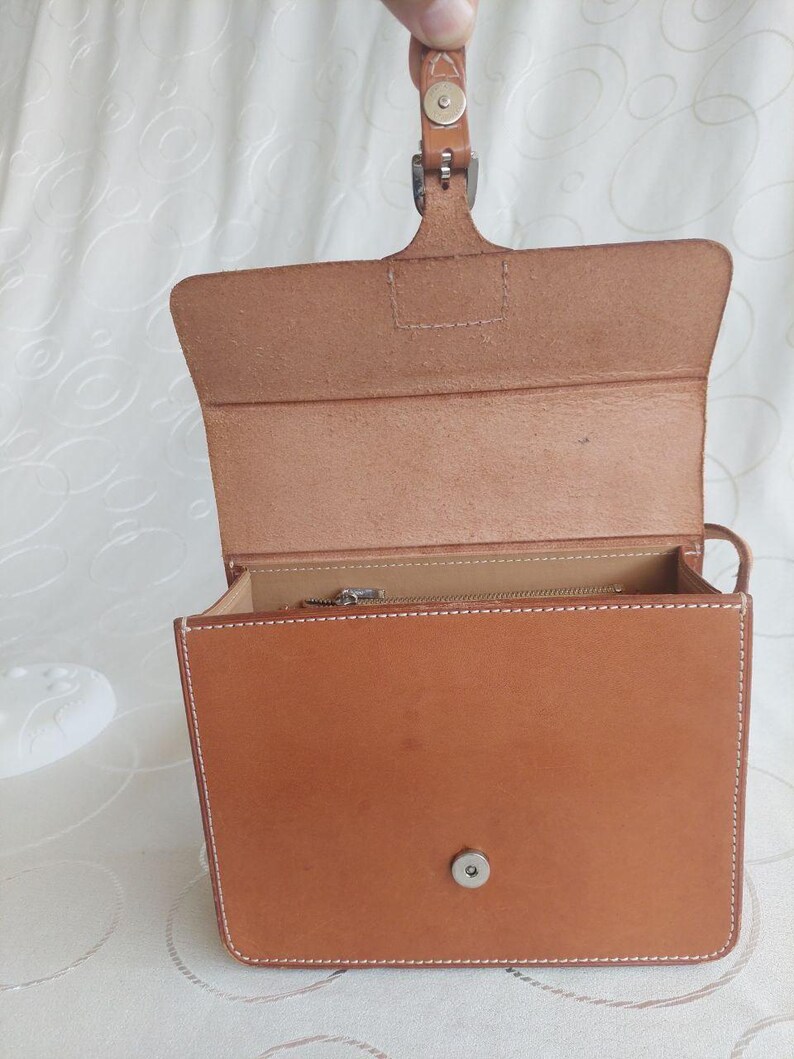 May include: Open, light brown leather handbag with a rectangular shape. The bag features white stitching, a top handle, and a metal clasp closure. The interior reveals a zippered pocket.