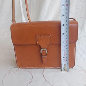May include: A rectangular, tan leather crossbody bag with a flap closure and a silver buckle. The bag has a shoulder strap and white stitching. The bag is approximately 8 inches tall.