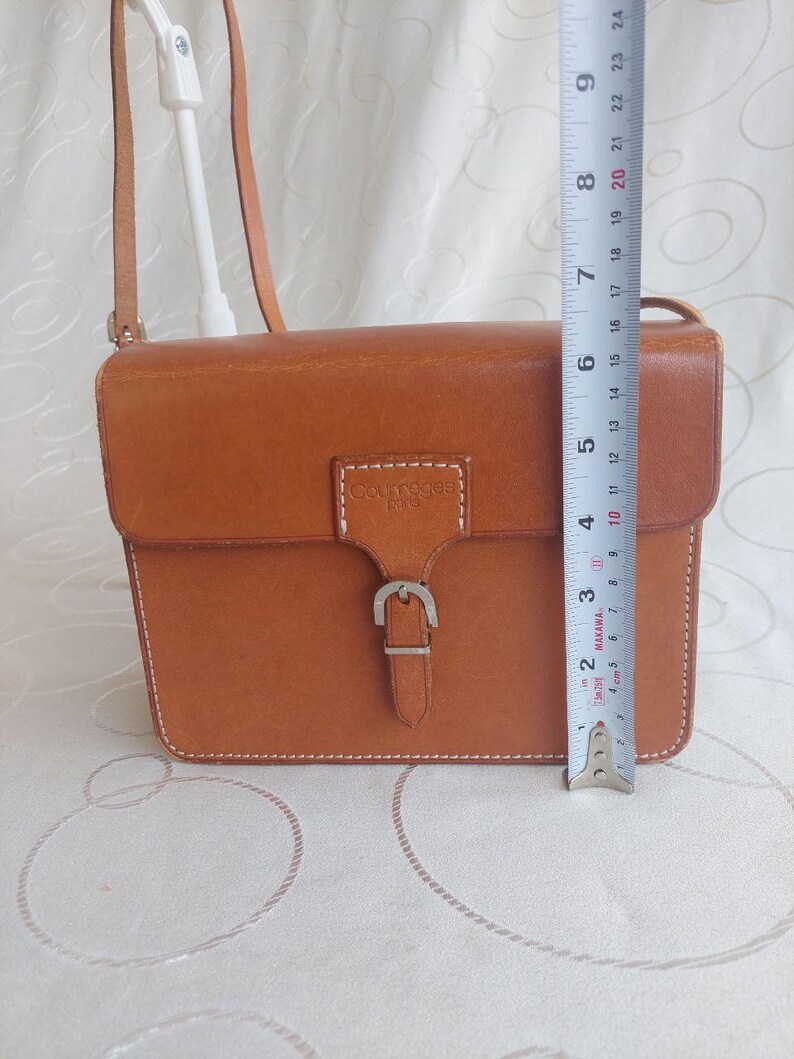 May include: A rectangular, tan leather crossbody bag with a flap closure and a silver buckle. The bag has a shoulder strap and white stitching. The bag is approximately 8 inches tall.