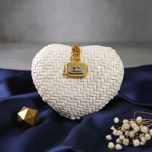 May include: A heart-shaped white woven handbag featuring a gold-tone chain strap attachment and a rectangular metallic clasp centered on the front. The clasp displays a silver-tone logo on a gold-tone frame, creating a distinct contrast.