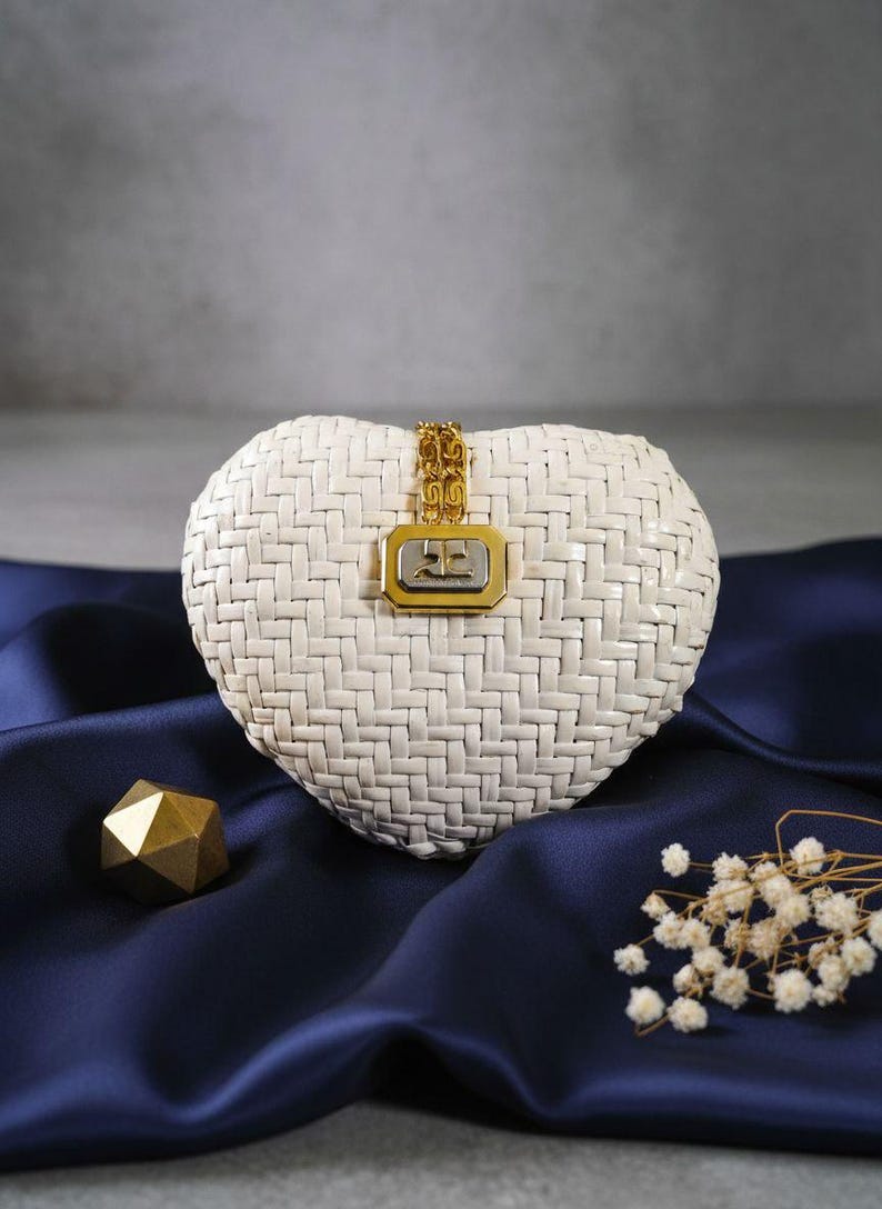 May include: A heart-shaped white woven handbag featuring a gold-tone chain strap attachment and a rectangular metallic clasp centered on the front. The clasp displays a silver-tone logo on a gold-tone frame, creating a distinct contrast.