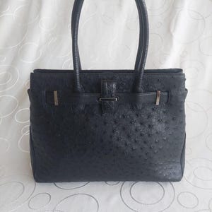 Op de afbeelding: Zwarte leren tote bag met een getextureerde afwerking en twee handvatten aan de bovenkant. De tas heeft een klepsluiting met een zilveren gesp en riem. Een klassieke stijl, geschikt voor dagelijks gebruik. Afmetingen: ongeveer 30 cm hoog en 40 cm breed.
